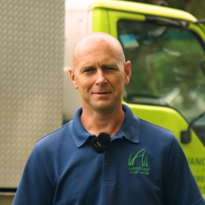 Andrew Powell (Founder of Advanced Turf Care) wearing a navy blue polo shirt with the logo "Advanced Turf Care" stands outdoors, smiling and looking directly at the camera. In the background, a green service vehicle is partially visible.