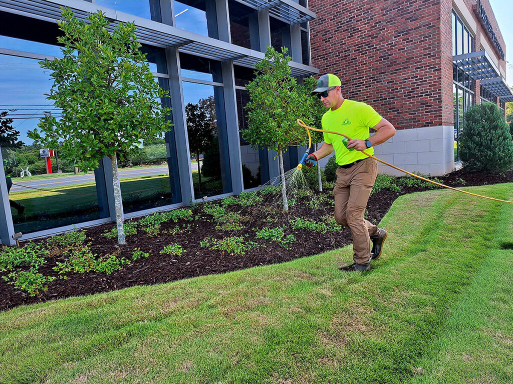 Andrew Powell (Founder of Advanced Turf Care) wearing a bright yellow shirt and a cap walks across a well-maintained green lawn, using a tool to tend to the flower beds surrounding small trees and shrubs near a modern red-brick building. The sky is clear.