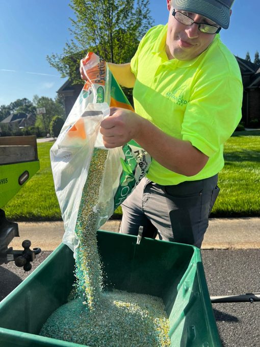 Advanced Turf Care Team member is pouring a bag of landscape material into a green container. The background features a well-maintained lawn and a clear blue sky on a sunny day.