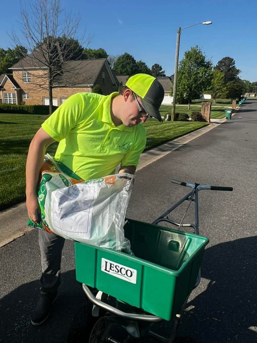 A young man from Advanced Turf Care in a bright yellow shirt and cap is preparing to use a green LESCO spreader. He is holding a bag of material, focusing intently on his task, while standing on a suburban street with neat houses and trees in the background.